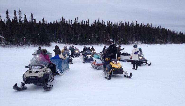 A group of people stand among snowmachines and boxes being towed on a snow covered area. 