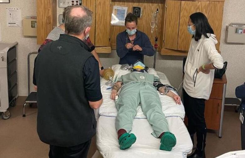 A group of people gather in a small room around a person lying on a hospital bed. 