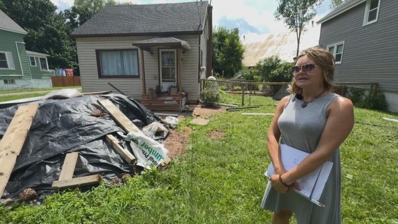 A woman in a grey dress and sunglasses stands on her property. Her home and property are being repaired. 