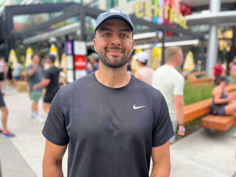 A man wearing a black Nike t-shirt and blue baseball cap stands in front of a Calgary pub.