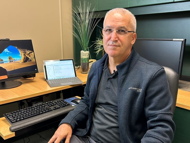 Man with some white hair sitting in an office wearing a blazer. He is front of a computer and a laptop.