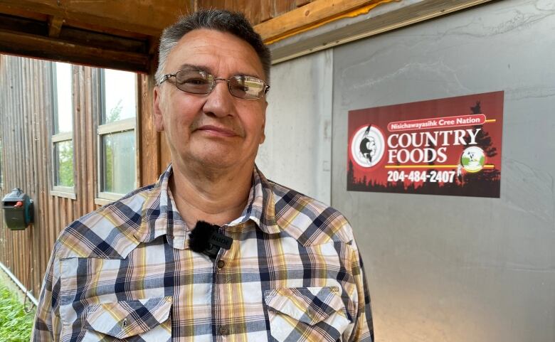 A man in a plaid shirt stands outside a building that has a sign reading 