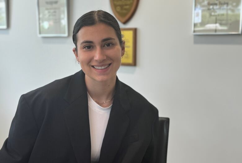 Realtor Anya Ettinger poses in a white shirt and black blazer in front of some diplomas on the wall.