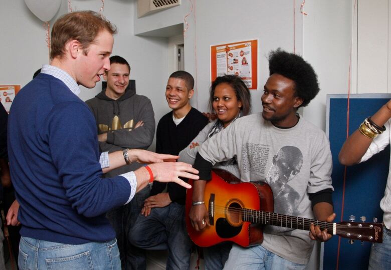 A person talks with other people, including one holding a guitarl.