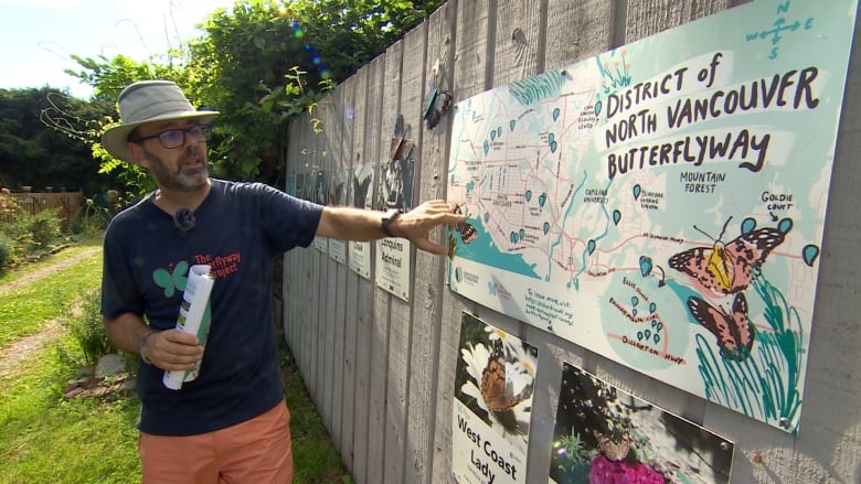 A man in a wide-brimmed hat stands outdoor along a fence. He points to a poster on the fence indicating butterfly habitat in the region.