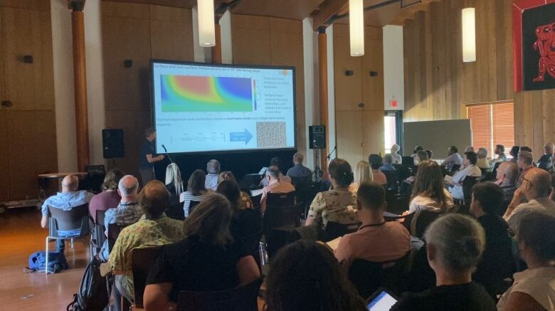 An audience in a large room watches a screen.