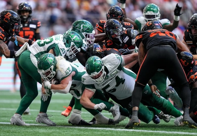 A mass of football players is seen, with one player diving over the outstretched leg of another.