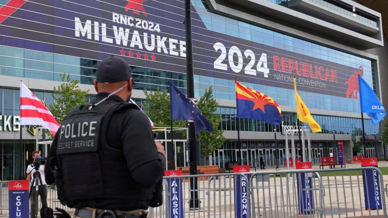 A man in a baseballcap and uniform with a visit that says 'Police Secret Service' stands with his back to the camera, looking toward steel barricades and a building that says 2024 Republican National Convention.