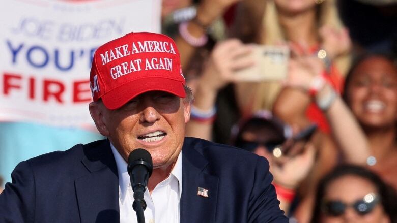 A man in a red baseball cap is seen squinting in an outdoor photo as he speaks at a podium. Onlookers are shown behind him.