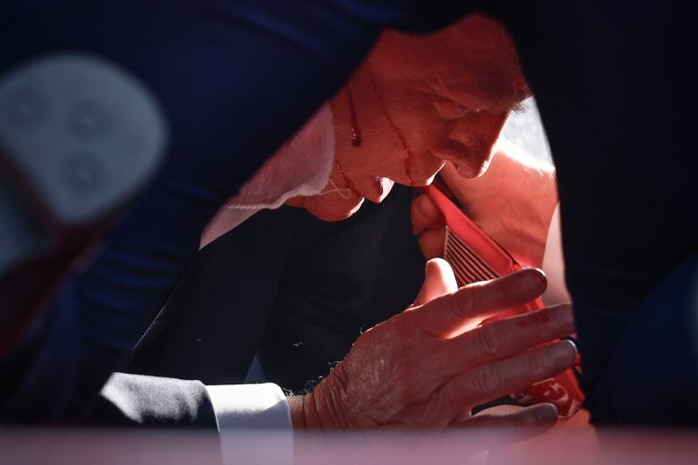 A man is just visible in extreme closeup lying on the ground, with a trickle of blood on his face and his hand visible.