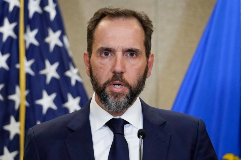 A bearded man wearing a suit and tie looks into the camera while speaking on a stage.