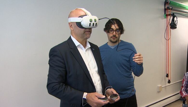 A white, bald man in a navy suit and white shirt wears a pair of white VR goggles with a wire trailing out the front being held by another man