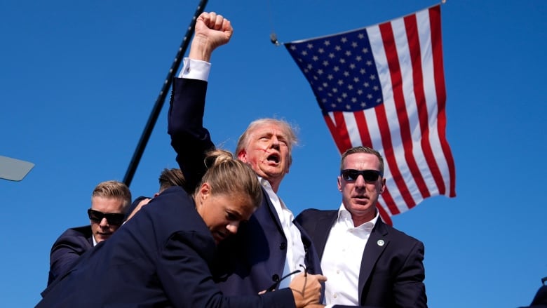 A man raises his fist with blood smeared across his face after being hit with a bullet on his ear with an American flag in the background.