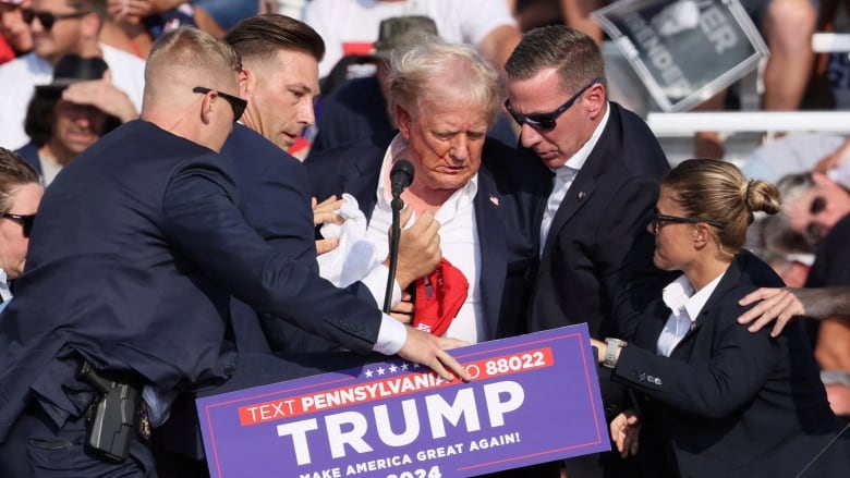 A man stumbles and is assisted by security personnel while bleeding from his ear on stage.