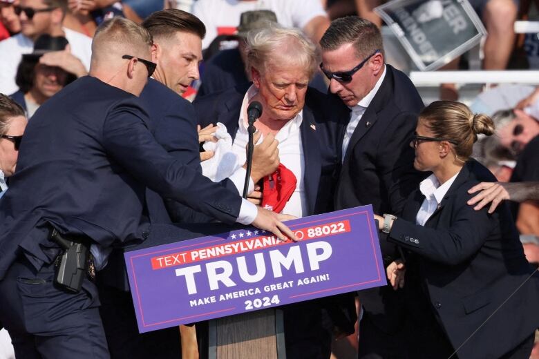 A man stumbles and is assisted by security personnel while bleeding from his ear on stage.