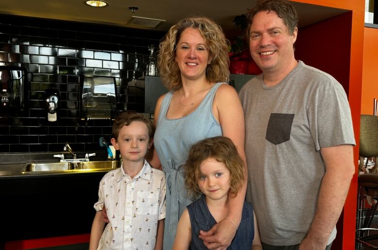 A woman in a blue shirt and curly blonde hair holds onto her two young children. A man in a grey t-shirt stands beside them smiling.