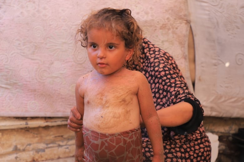 A little girl stands in a tent in red shorts and has sand rubbed on her 