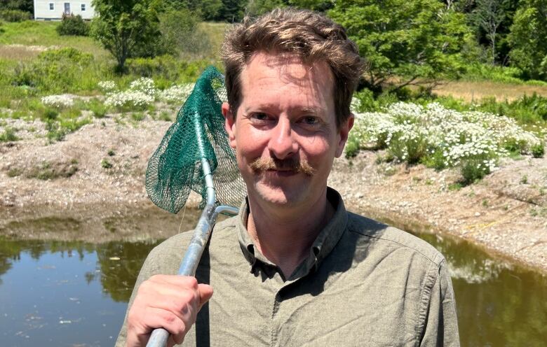 A white man with curly brown hair and moustache. Holding a frog catching net in his hand, resting it on his shoulder.