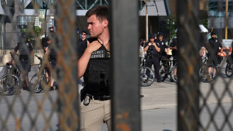 A member of the Secret Service holds onto the collar of their bulletproof vest as they stand behind a metal fence.