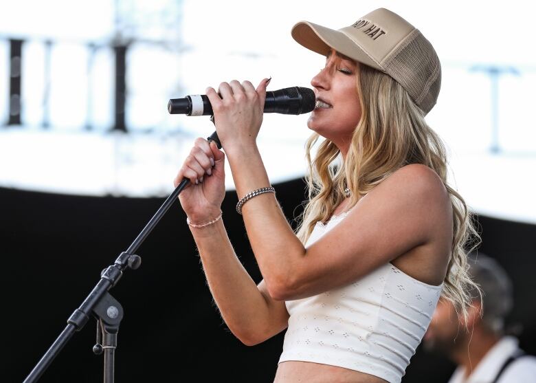 singer ingrid andress performs in tan hat and white tank top, singing into a microphone on a stand