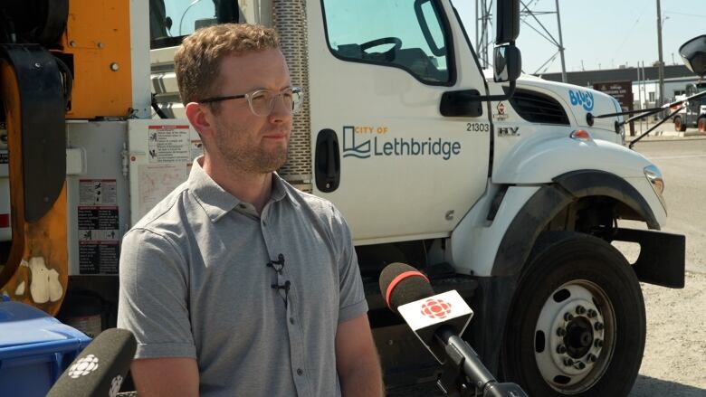 A man wearing glasses stands in front of a white truck with city of lethbridge on the door and microphones in front of him