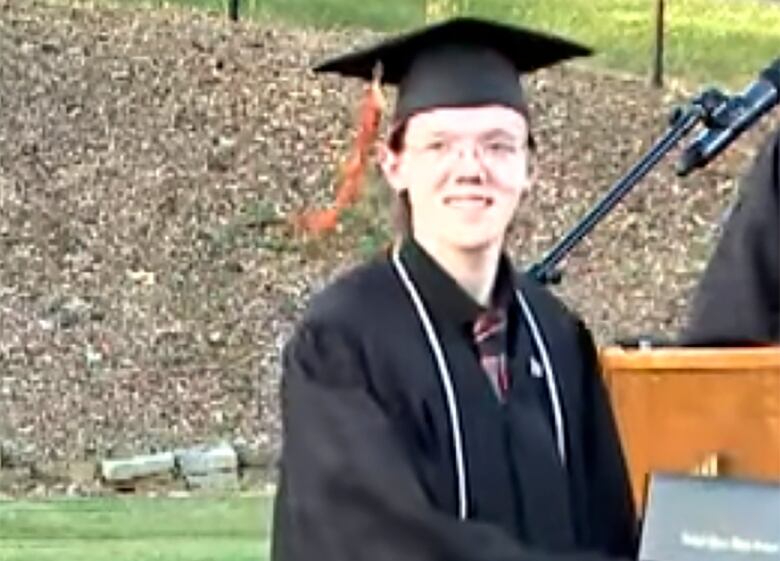 A boy wearing glasses and a dark, graduation-style cap and gown is shown in an outdoor setting.