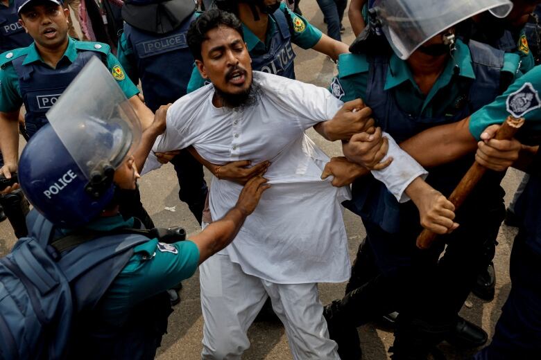 Police in full riot gear wrestle with a man wearing white clothing.