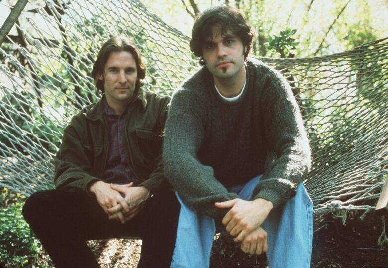 Two men in their late 20s sit on a hammock, facing the camera.