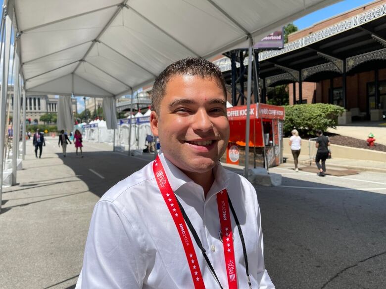 A young man wearing a white shirt with a red lanyard stands under a large tent.