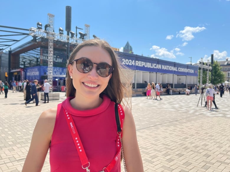 A young woman wearing a pink dress, black sunglasses and a red lanyard stands in a plaza.