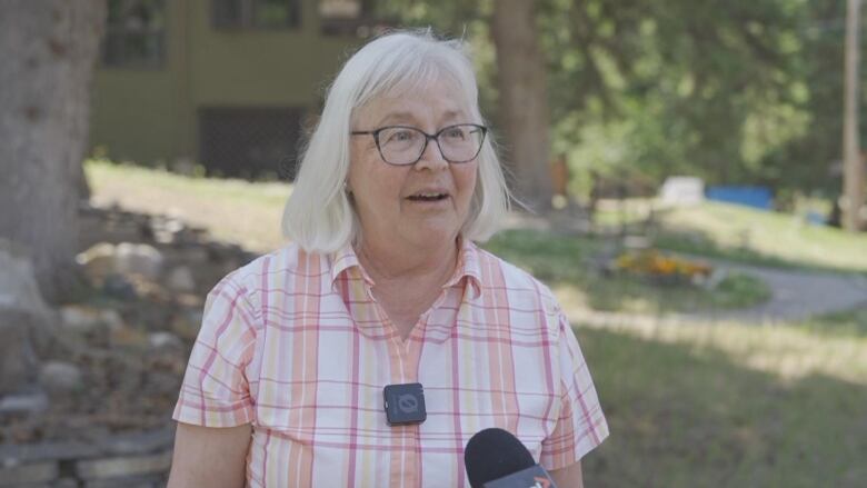 a woman with glasses speaks into a microphone. there are trees and grass behind her.