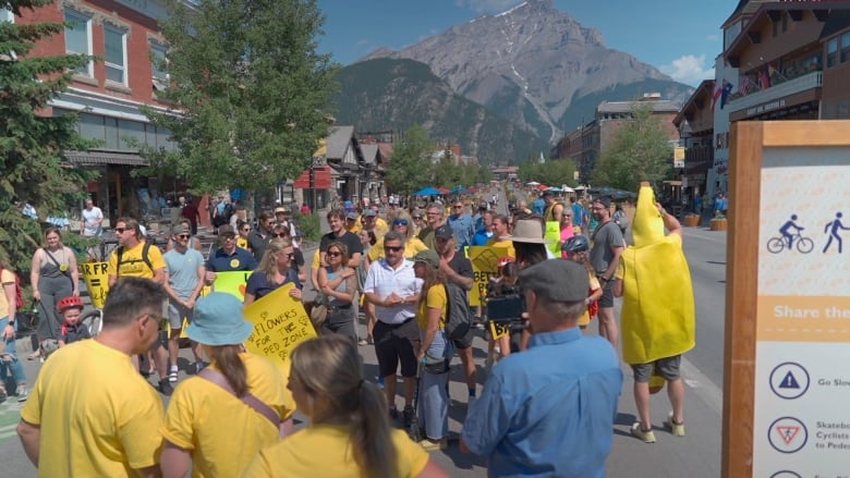 a group of people in yellow shirts stand on a street holding yellow signs. there is a large mountain in the background.