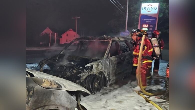 A burnt car is shown destroyed in the darkness of night with a firefighter standing next to it in red firefighting gear.