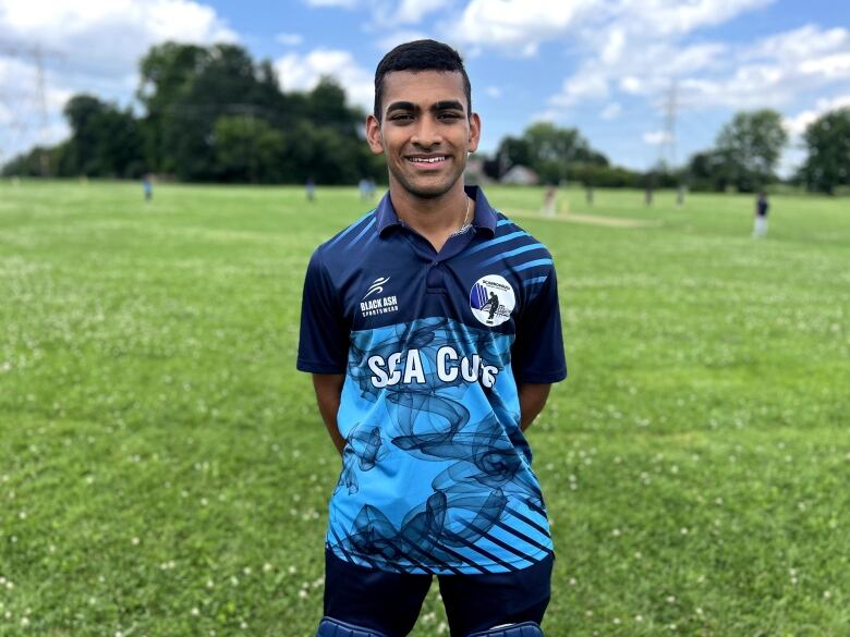 A young brown man smiles for a portrait on a grass cricket field. 