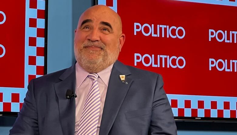 A bald man in a suit and tie smiles as he stands against a red background featuring a sign reading 'Politico.' 