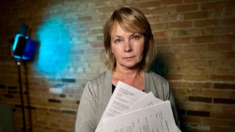 A woman wearing a grey sweater is standing in front of a brick wall holding documents printed on white paper. 