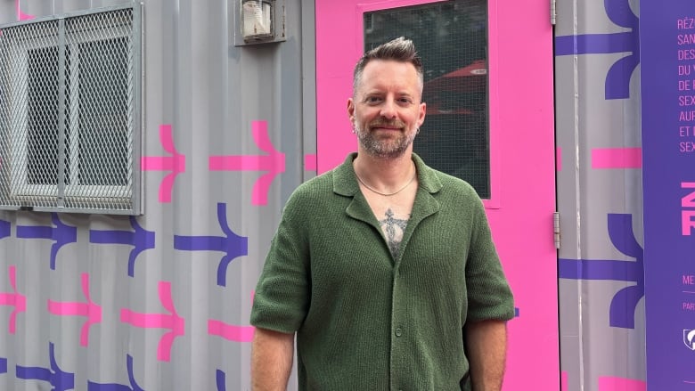 A man in green cotton sweater standing outside in front of a shipping container that's been converted 