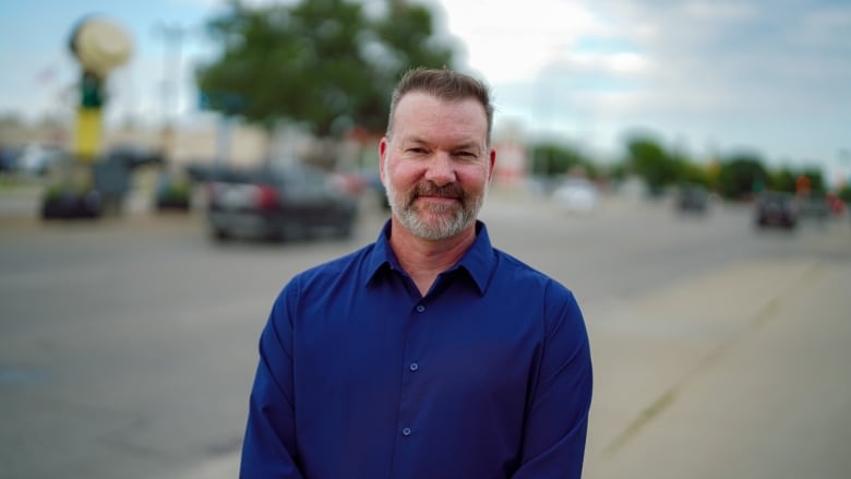 A man in a blue shirt stands on a sidewalk, next to a highway in Transcona.