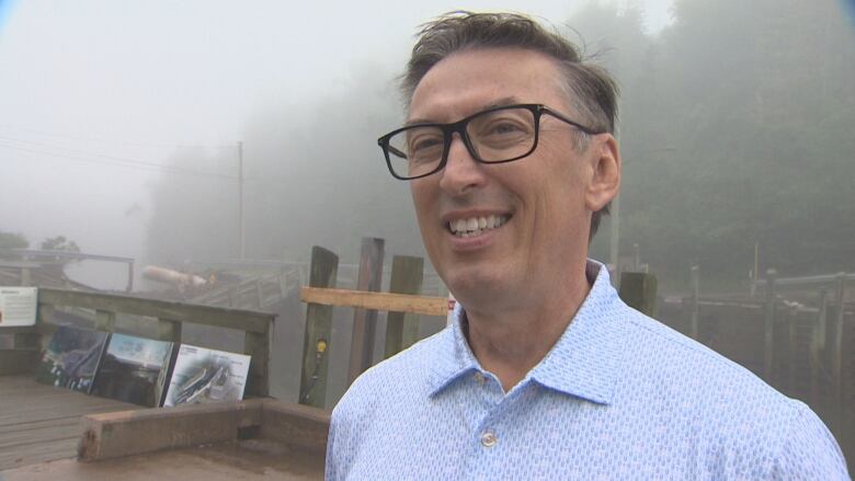 Man in glasses and blue shirt stands outdoors on a foggy day.