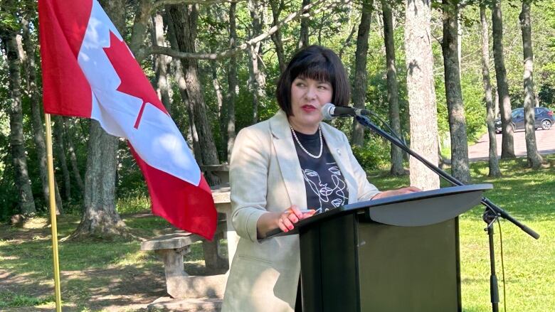 A white lady with black hair, standing at a podium in a park.