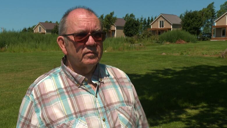 A man with a plaid shirt and sunglasses stands in front of summer cottages.