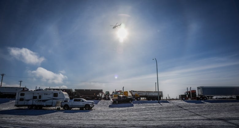 Several trucks and trailers block a snowy highway on a sunny day as a helicopter flies overhead.