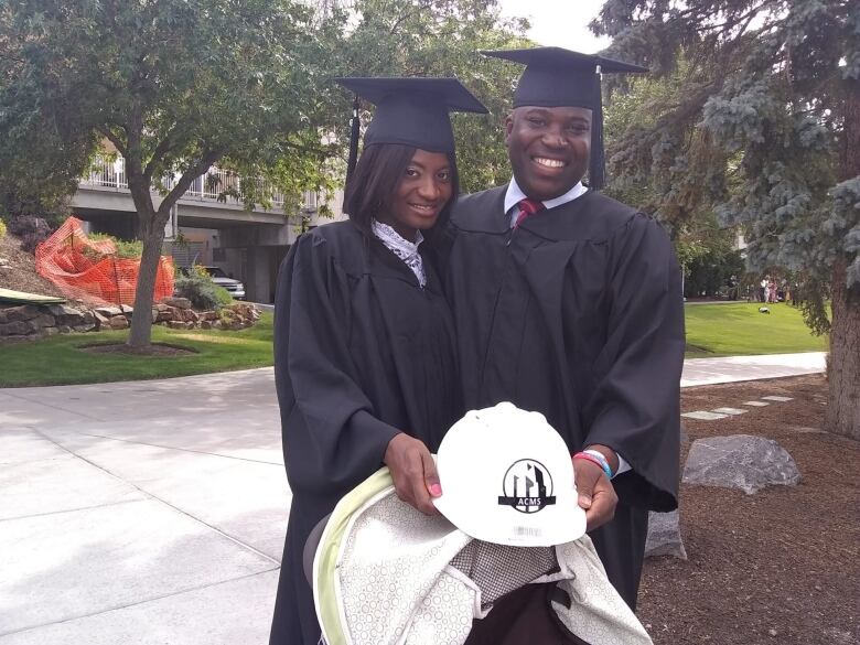 A woman and man in graduation caps and gowns holding a construction hat between them. 