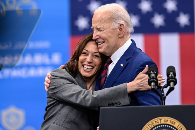 Two people smile while they embrace behind a lectern.