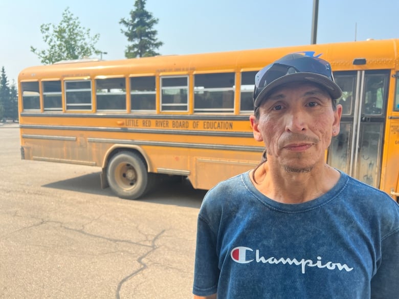 An Indigenous man, wearing a ball cap with sunglasses resting on the brim and a blue t-shirt, is standing next to a yellow school bus. His expression is neutral.