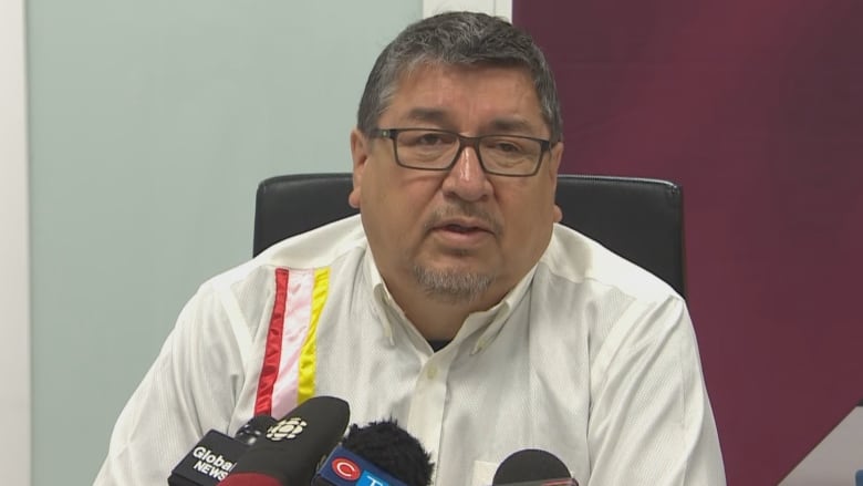 A man with short grey hair sits behind microphones at a table.