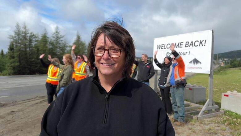 A smiling woman wearing a black sweater stands in front of a group of waving people on the side of a road.