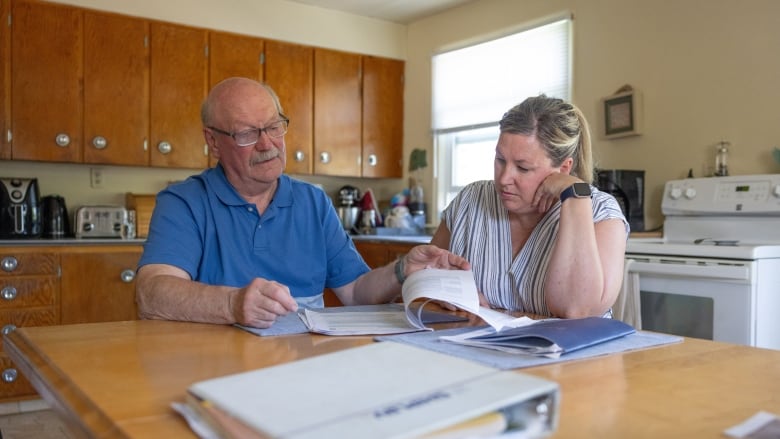 Two people sit at a kitchen table looking at a paper contract between them. One is an older Caucasian man, balding and wearing glasses and a blue shirt. The other person is a Caucasian woman with blonde hair, wearing a white and blue striped dress. 