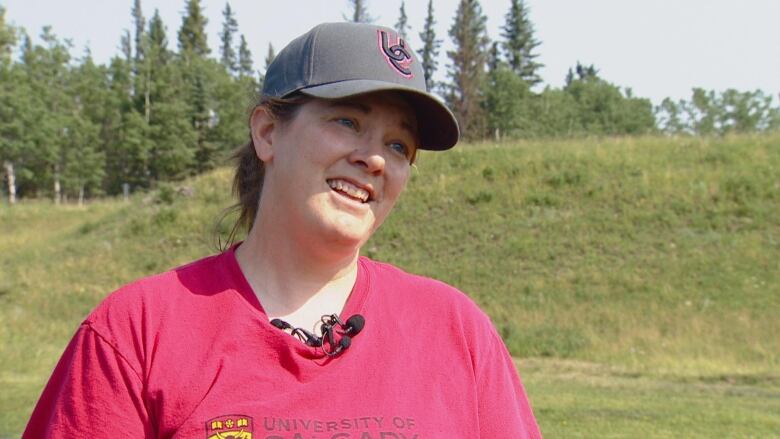 A close-up shot of a woman in a red University of Calgary T-shirt. She is seen wearing a cap and is smiling.