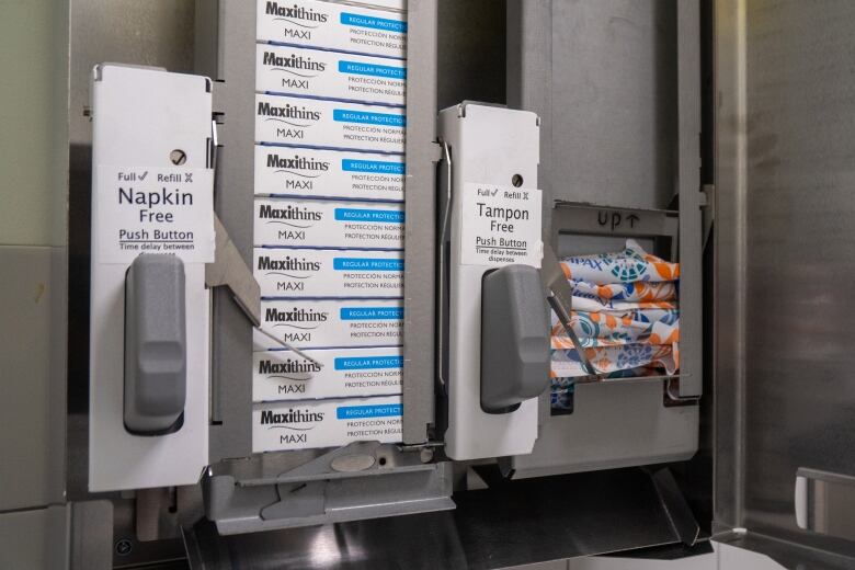 A tampon and pad dispenser is seen in a washroon, filled with packages of tampons and small boxes of pads.